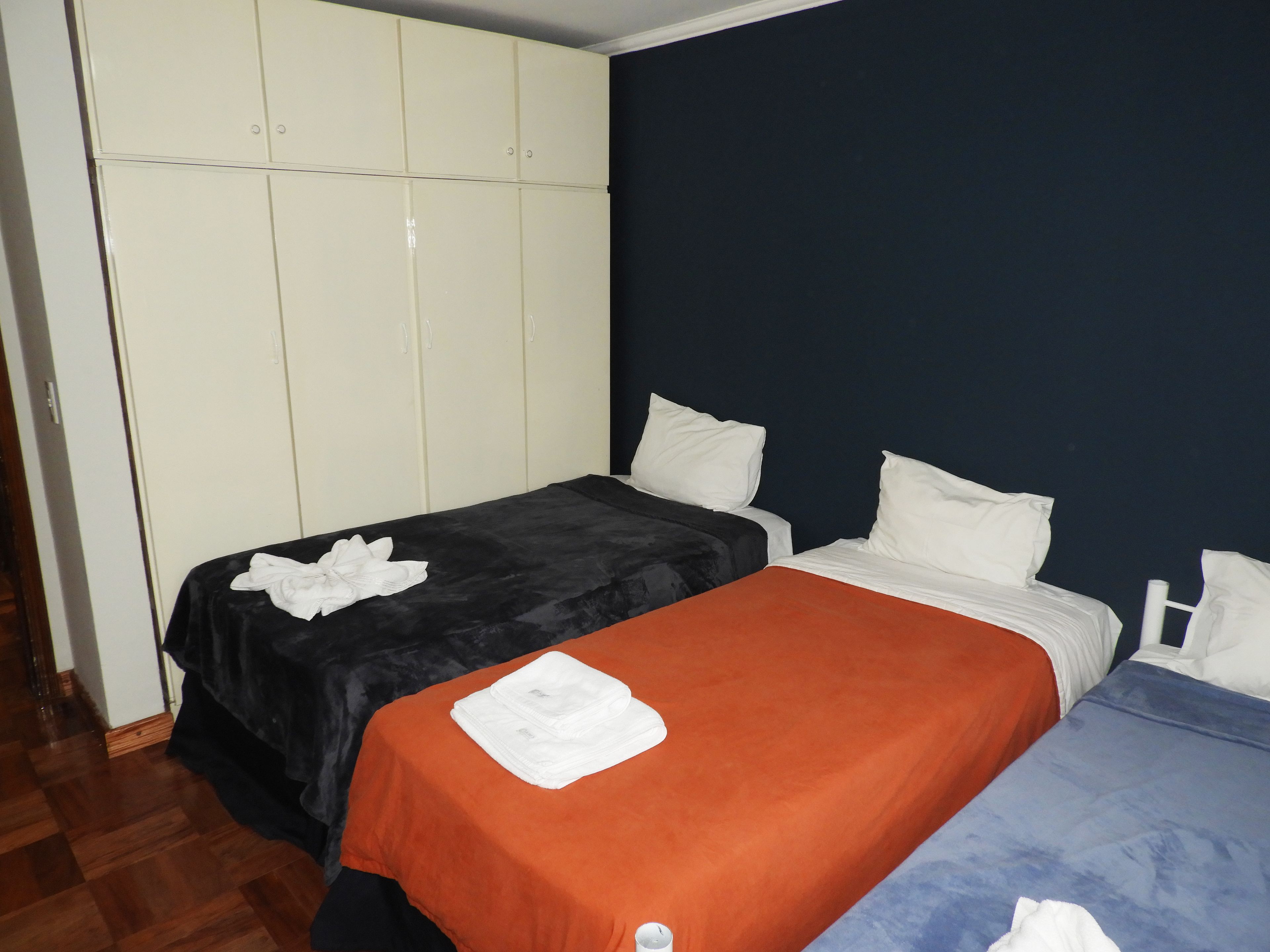 Una habitación con dos camas, una con una colcha naranja y la otra con una colcha azul. Se observa un armario blanco con múltiples puertas y una pared azul oscuro.