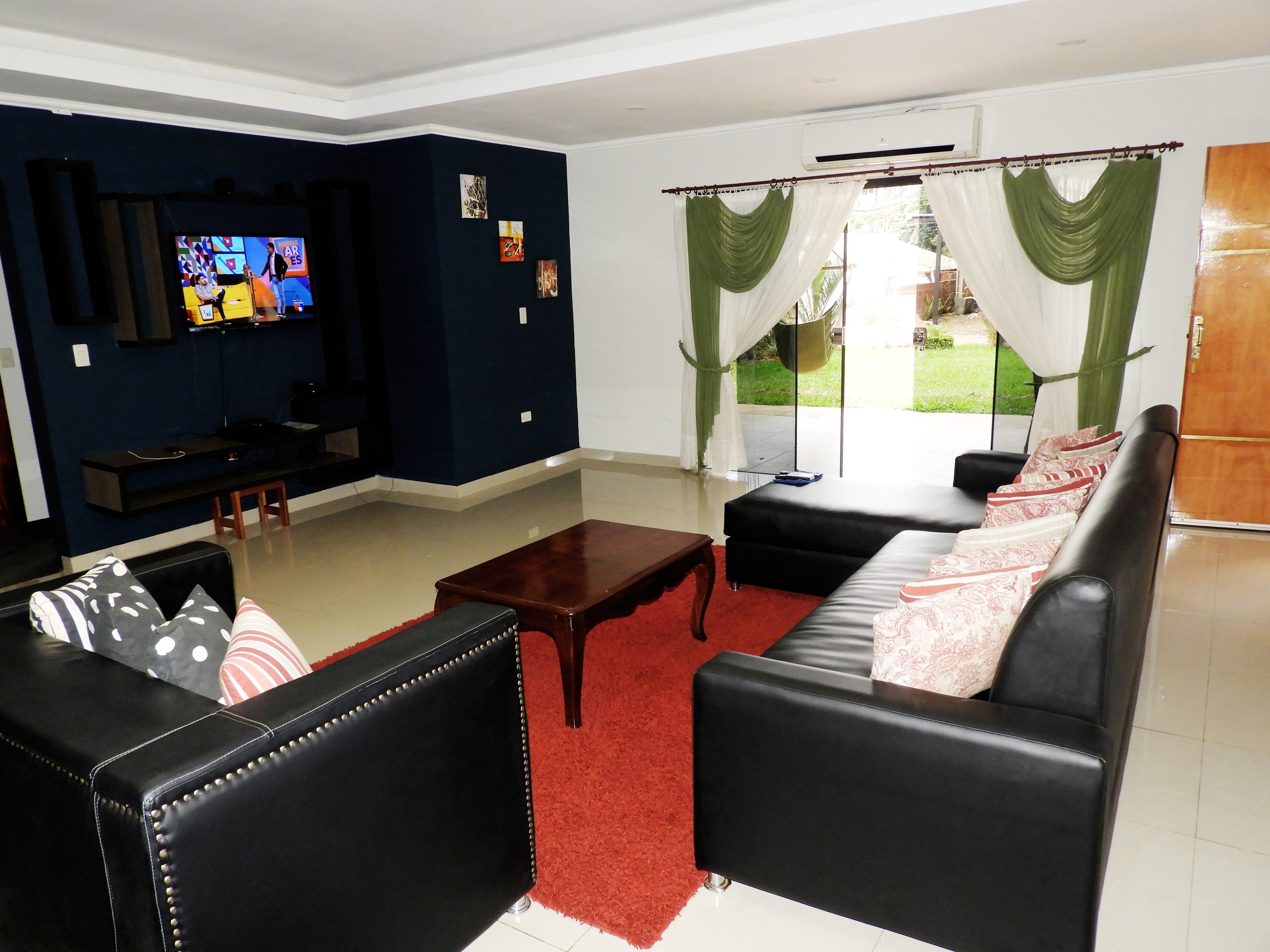 A living room with a black sofa, a coffee table, and a TV. There is a window with green curtains.