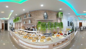 Uma praça de alimentação moderna e bem iluminada, com diversas opções de comida expostas em um balcão curvo, cercada por áreas de estar e plantas decorativas.