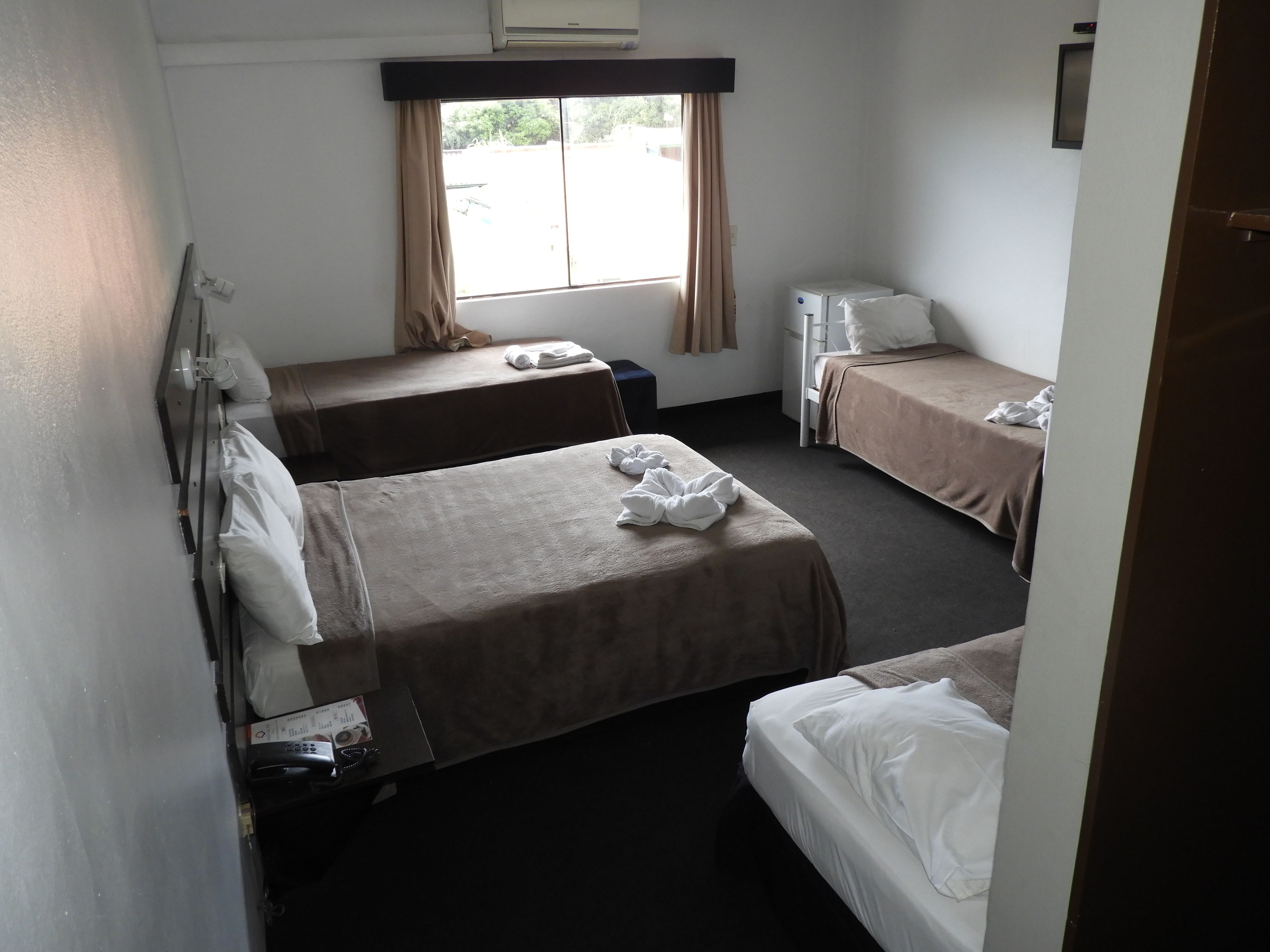 A hotel room with two single beds, a small desk, a chair, a window with curtains, and a wall-mounted air conditioner.
