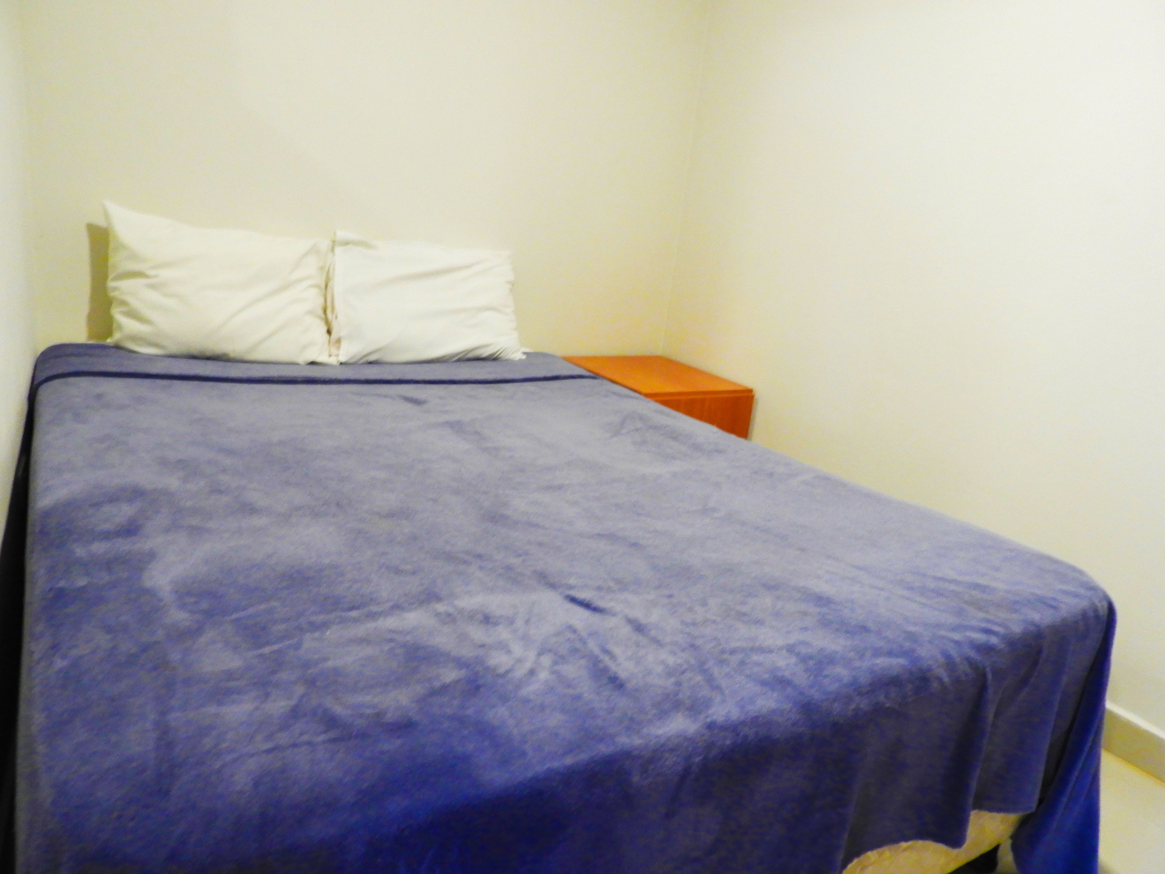 Una cama individual con colcha azul y almohada blanca en una habitación con pared blanca lisa y mesita de noche de madera.