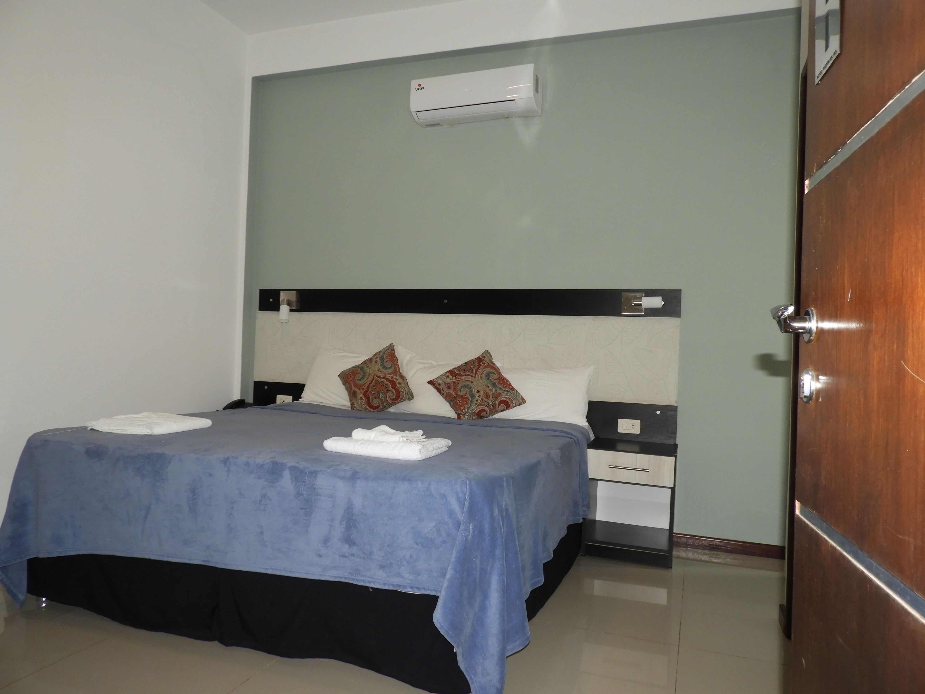 Una habitación de hotel con cama, mesita de noche, cajón, unidad de aire acondicionado y puerta de madera.