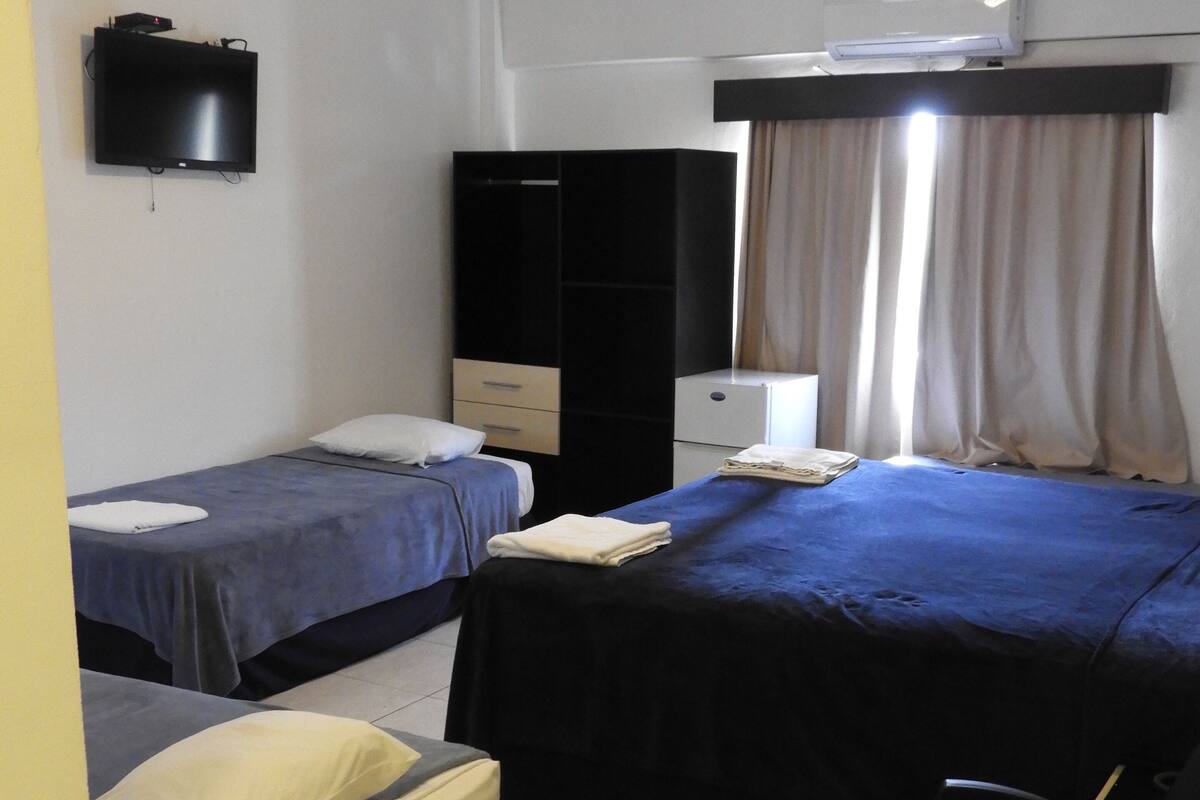 A room with two beds, a desk, a chair, a wardrobe, a TV, and an air conditioning unit.