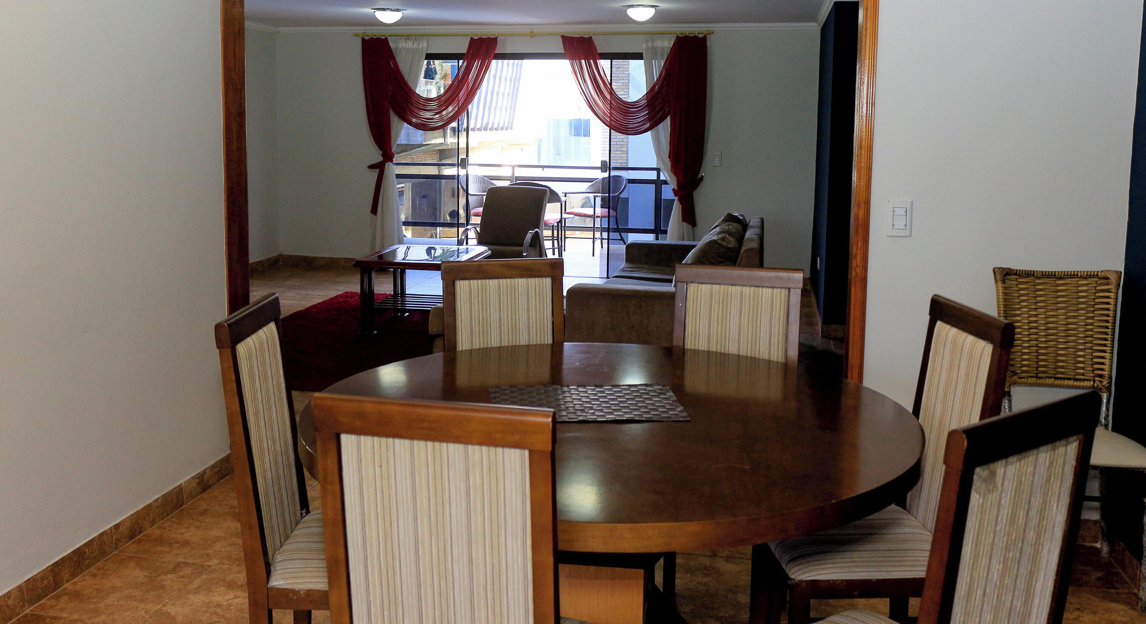 A dining area with a round table and six chairs, a view of the outside through a window with red curtains, and a balcony with chairs.