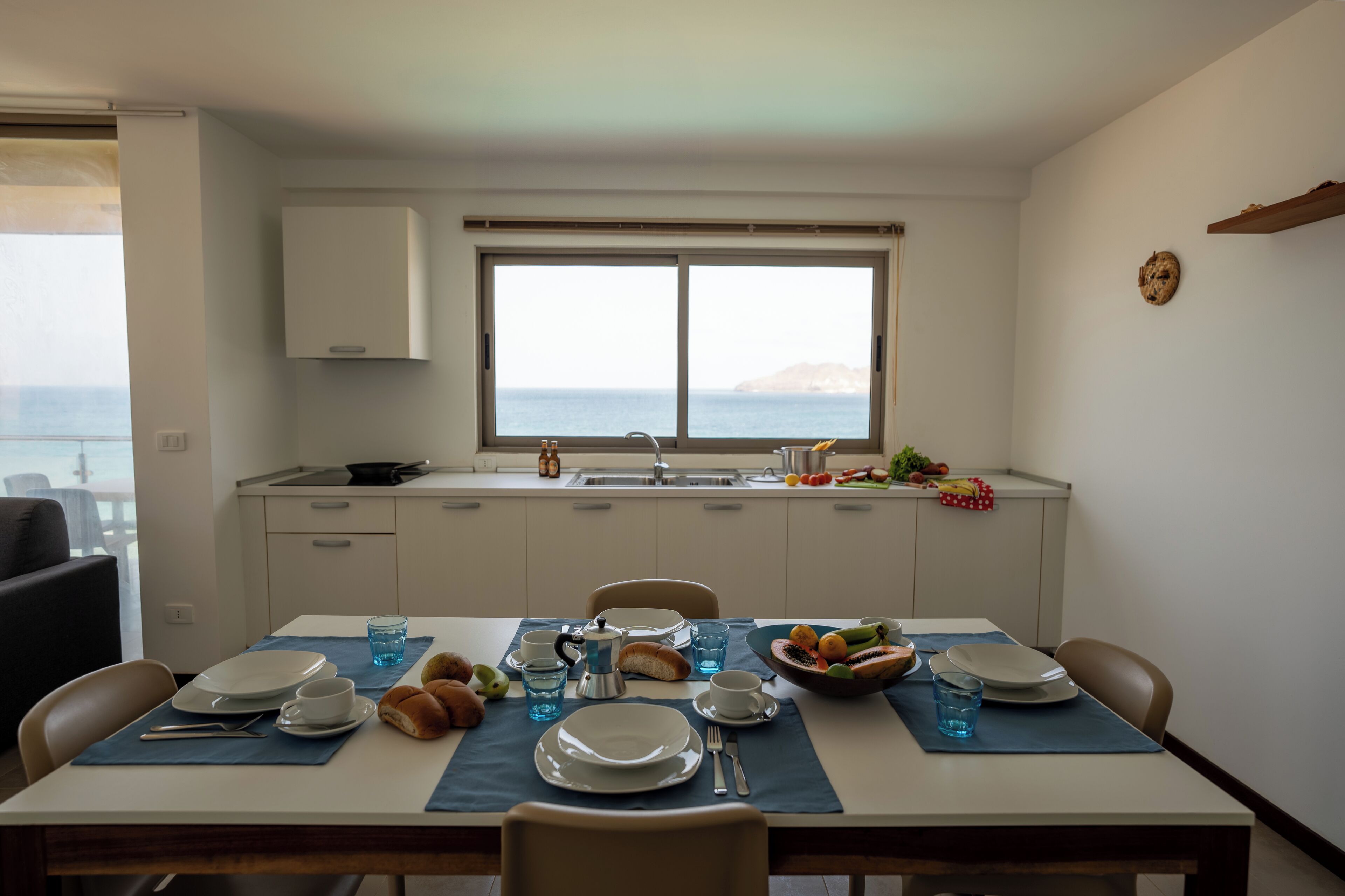 Un comedor con una mesa dispuesta para dos personas, una ventana con vistas al mar y una cocina al fondo.