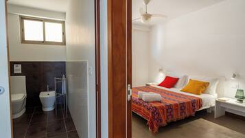 A hotel room with a bed, a nightstand, a bedside lamp, a window, a ceiling fan, and a bathroom with a toilet and bidet.