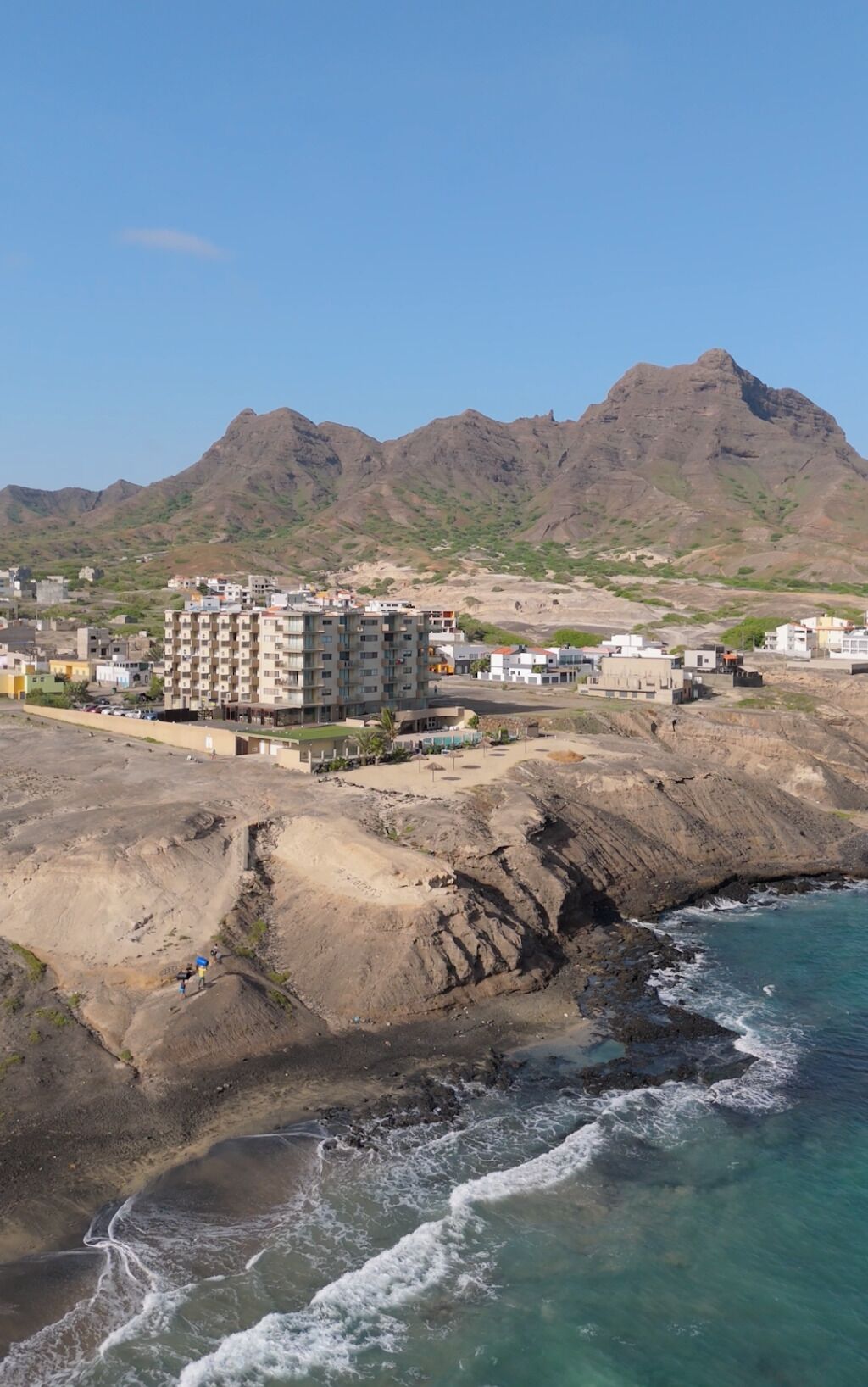 Een hotel aan de kust met een rotsachtige kustlijn, een berg op de achtergrond en een heldere lucht.