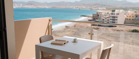 A balcony with a white table and chairs, overlooking a beach and cityscape.