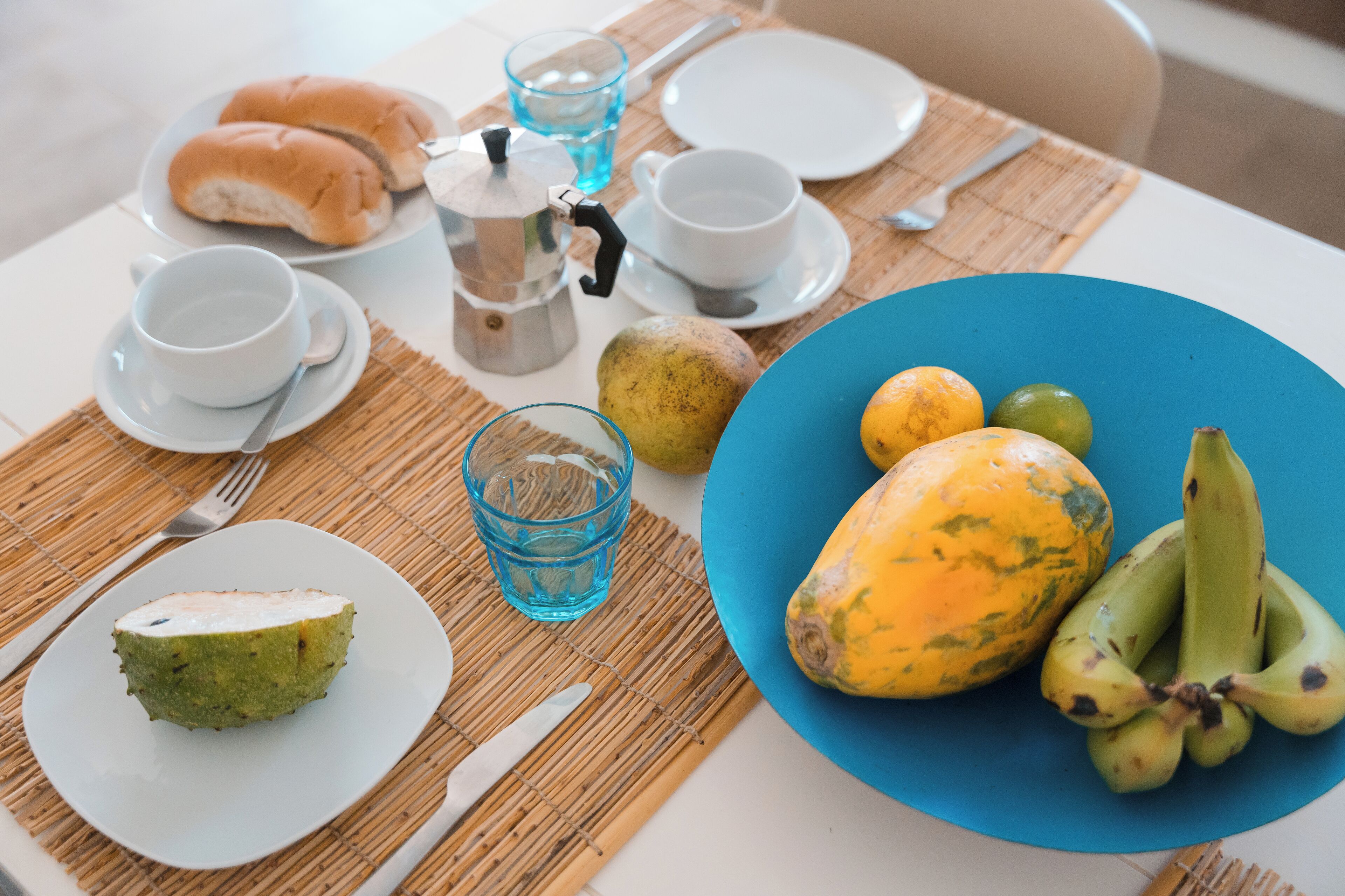 Una mesa de desayuno con platos de comida, tazas, una cafetera y un bol azul con frutas.