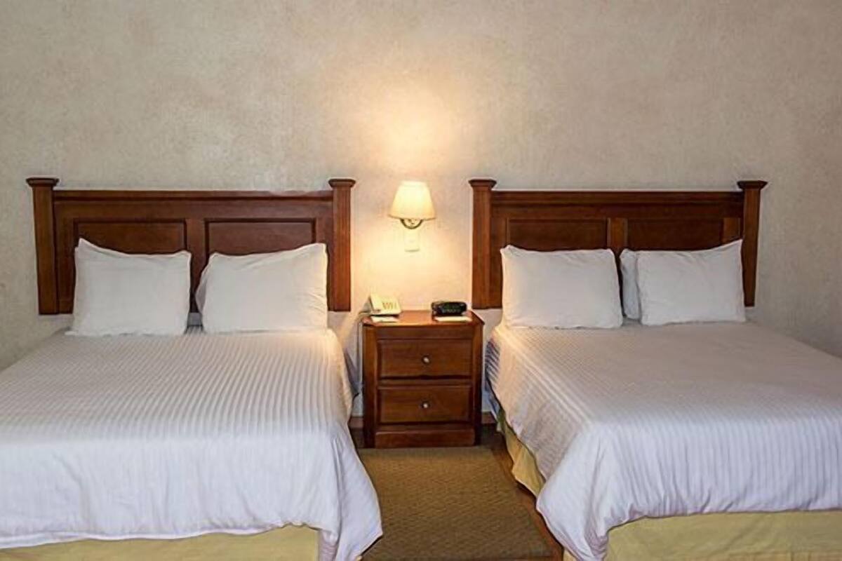 A hotel room with two single beds, wooden headboards, a nightstand with a lamp, and a small drawer.