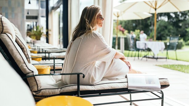 A person sitting on a cushioned bench with a book, in a well-lit indoor area with large windows and outdoor seating visible. - Garden Hotel Reinhart (Prien am Chiemsee)