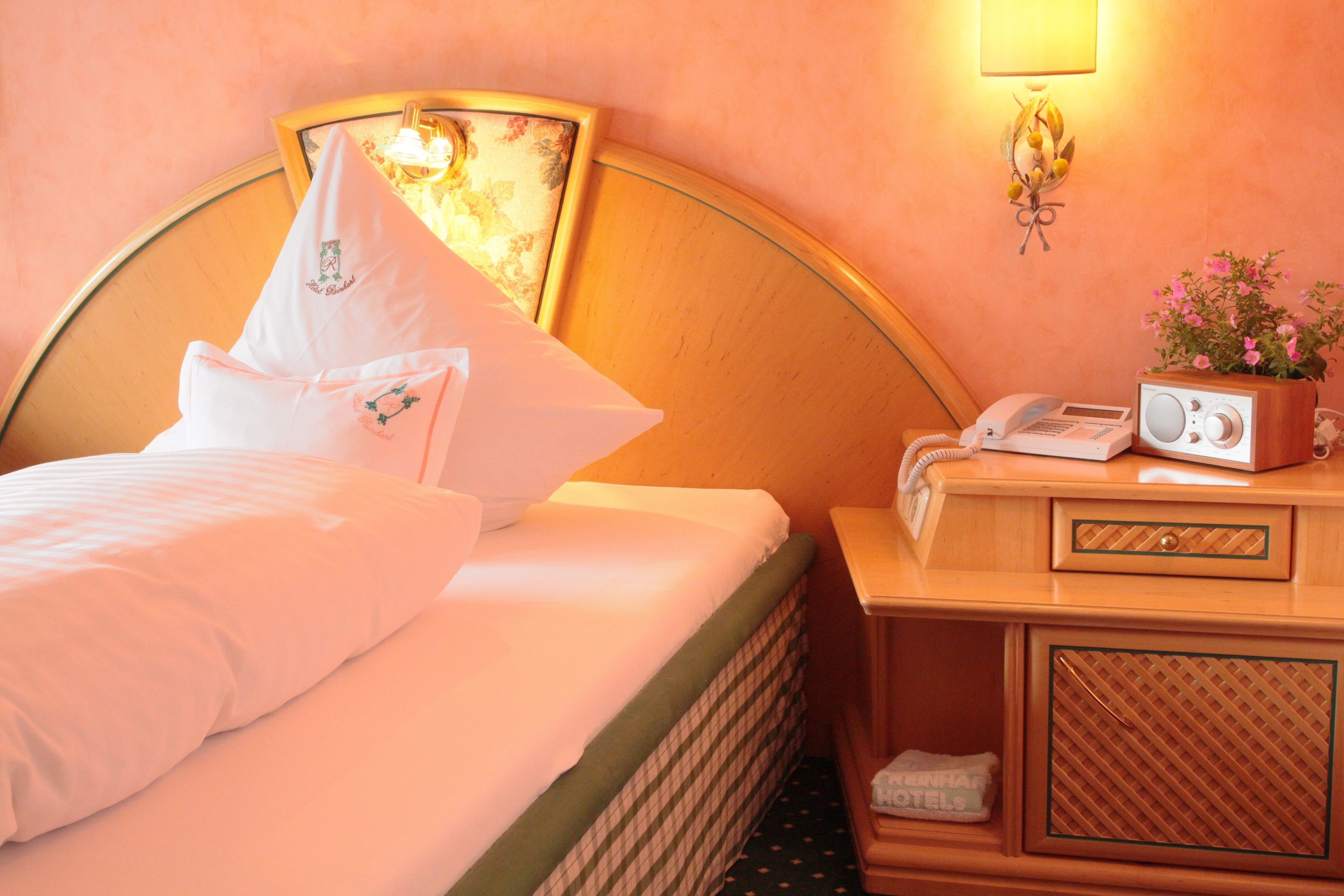 Quarto de hotel com cama, mesa de cabeceira com rádio e telefone, e luminária na parede.