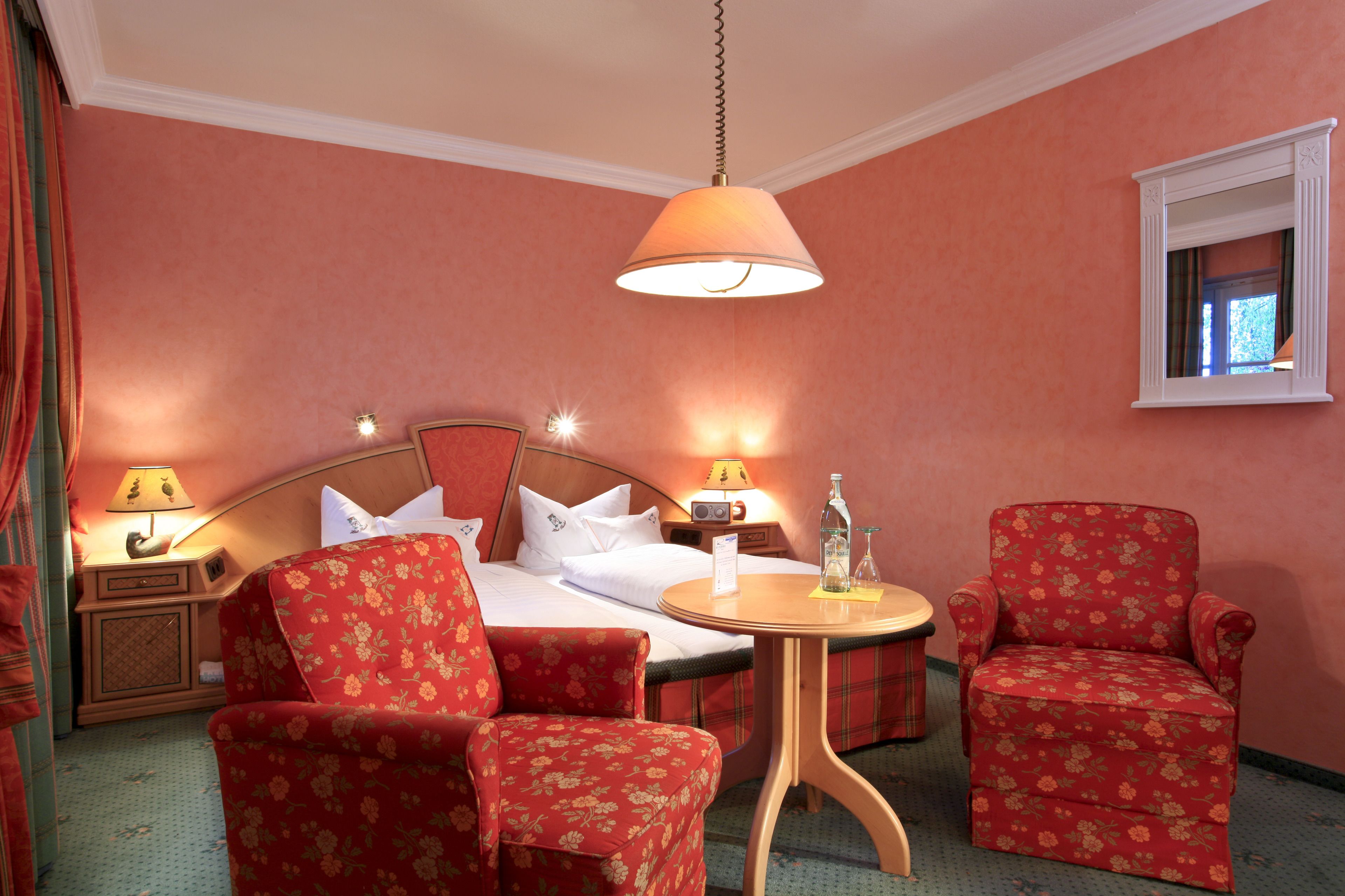 Une chambre d’hôtel avec un lit, deux fauteuils à motifs floraux, une table ronde en bois, une table de chevet avec une lampe et un miroir au mur.