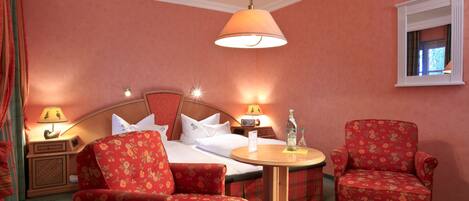 Une chambre d’hôtel avec un lit, deux fauteuils à motifs floraux, une table ronde en bois, une table de chevet avec une lampe et un miroir au mur.