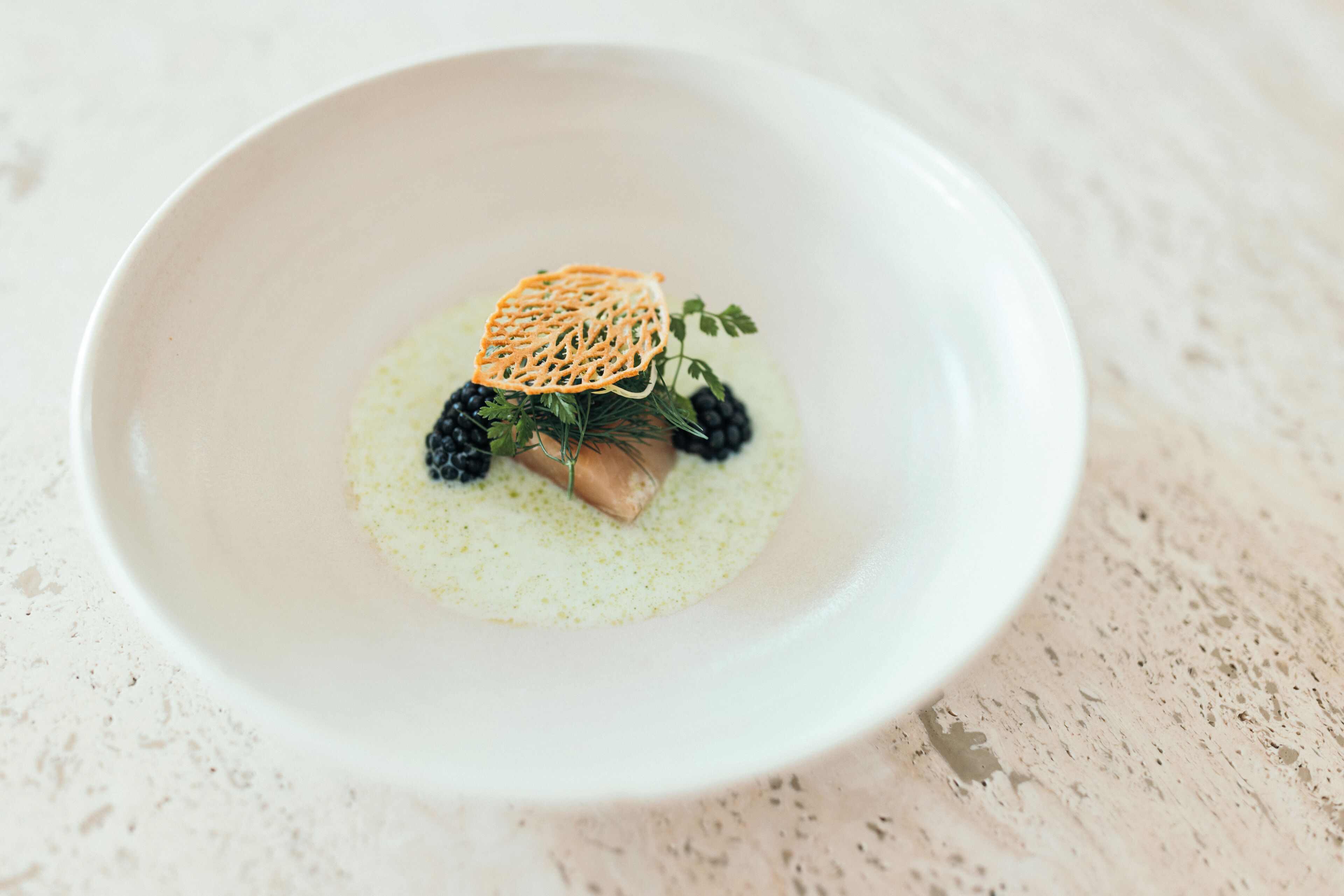 A white bowl with a dessert featuring a piece of cake, blackberries, and a decorative leaf on a speckled surface.