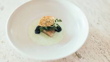 A white bowl with a dessert featuring a piece of cake, blackberries, and a decorative leaf on a speckled surface.