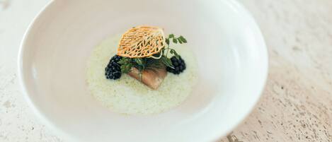 A white bowl with a dessert featuring a piece of cake, blackberries, and a decorative leaf on a speckled surface.