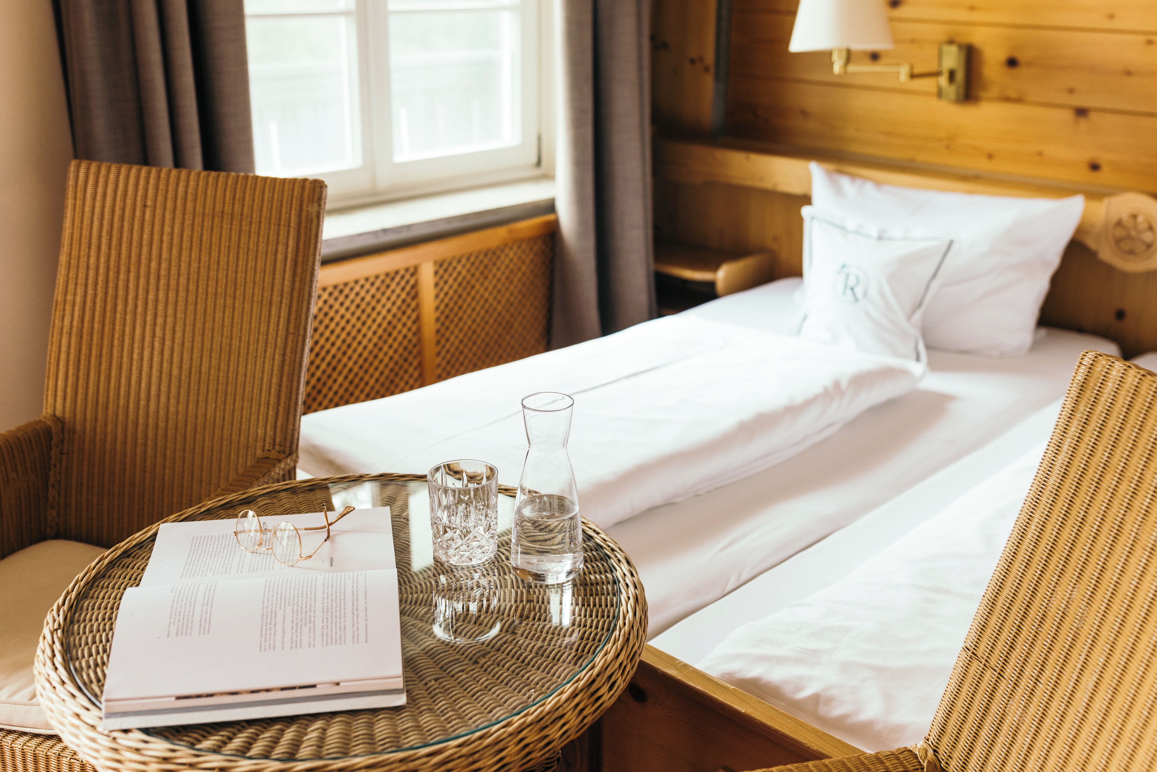 A neatly made bed with white linens, a wicker chair, a small round table with a glass of water, and a book.