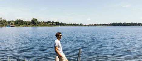 A person standing on a wooden dock extending into a lake, with a clear sky and distant trees.
