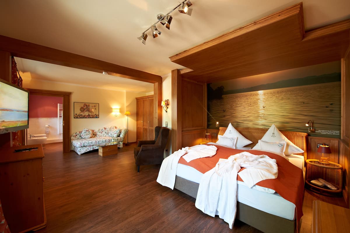 Habitación de hotel con piso de madera, cama con ropa de cama blanca, armario de madera, sillón pequeño y un cuadro en la pared.