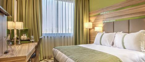 Habitación de hotel con una cama grande, un escritorio con silla, una lámpara y vistas a la ciudad a través de la ventana.