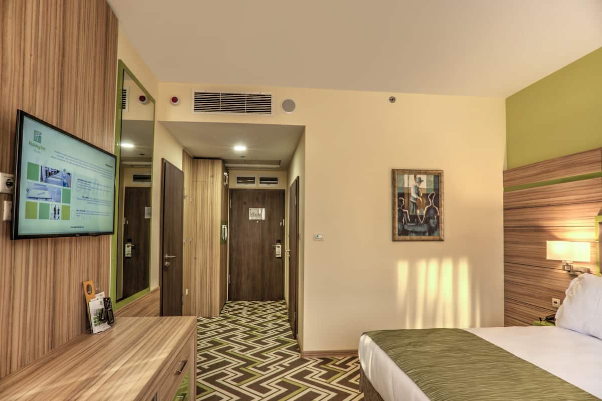 A hotel room with a bed, a TV mounted on the wall, a desk with a remote control, and a patterned floor.