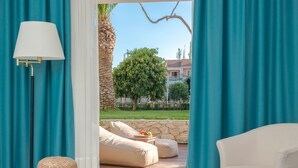 A room with a view of a garden, a lounge area with a table and chairs, and a large window with curtains. - Bitzaro Palace Hotel - All inclusive (Zakynthos)