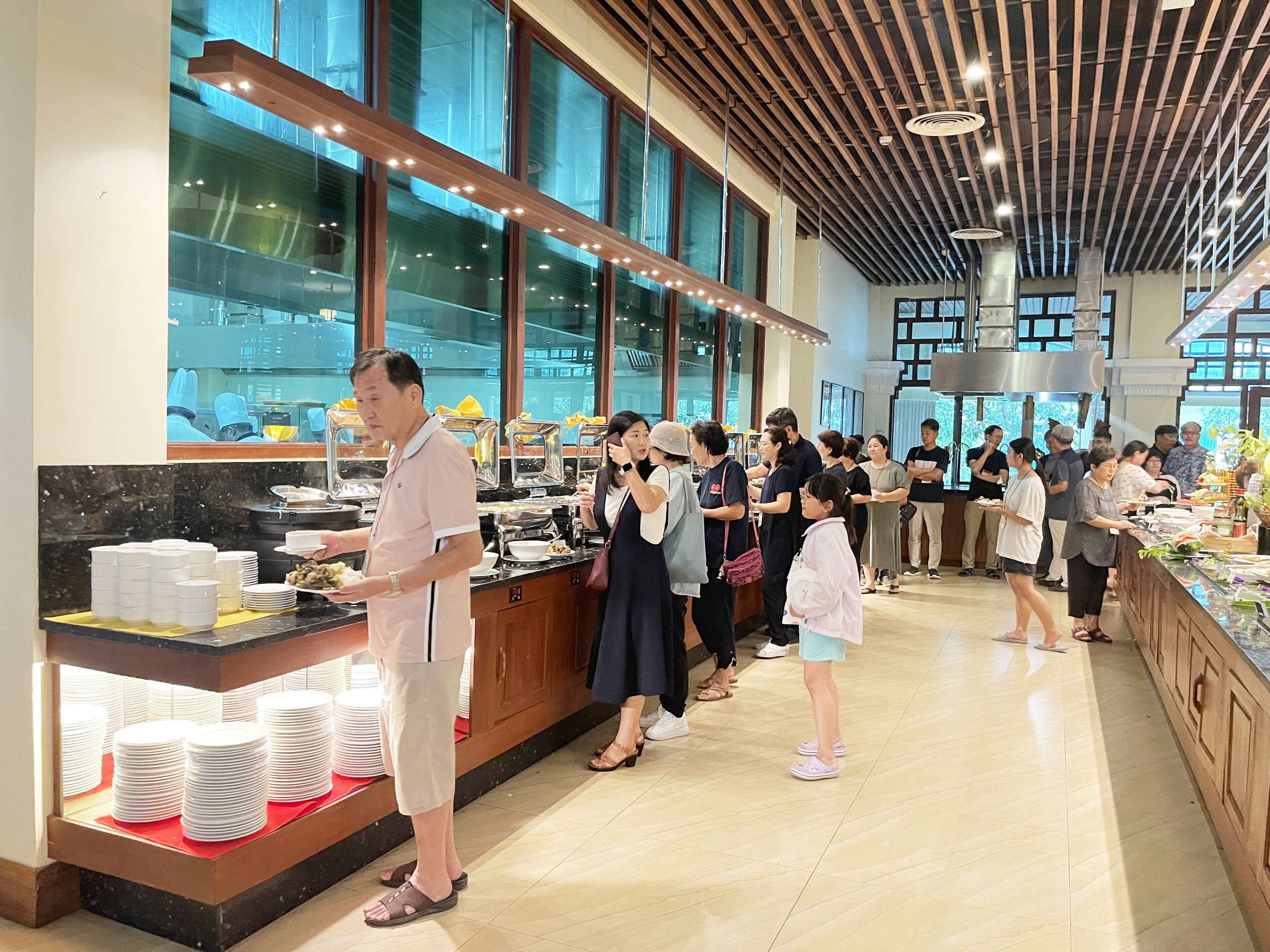 A buffet area with a counter displaying various dishes, a group of people queuing, and a modern interior with large windows and a wooden ceiling.