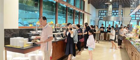 A buffet area with a counter displaying various dishes, a group of people queuing, and a modern interior with large windows and a wooden ceiling.