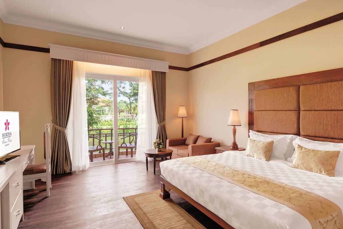 Ein Hotelzimmer mit einem großen Bett, einem Schreibtisch, einem Sessel, einer Couch und einem Balkon mit Vorhängen.