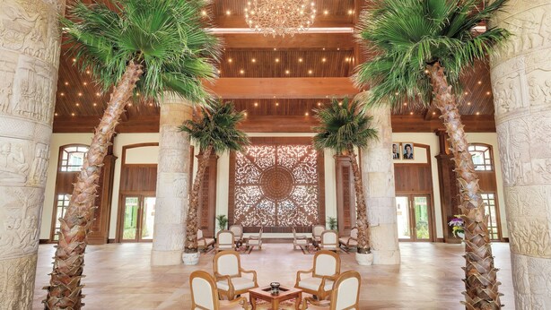 Een ruime lobby met hoog plafond, houten balken, een kroonluchter, palmbomen en een decoratieve wand.