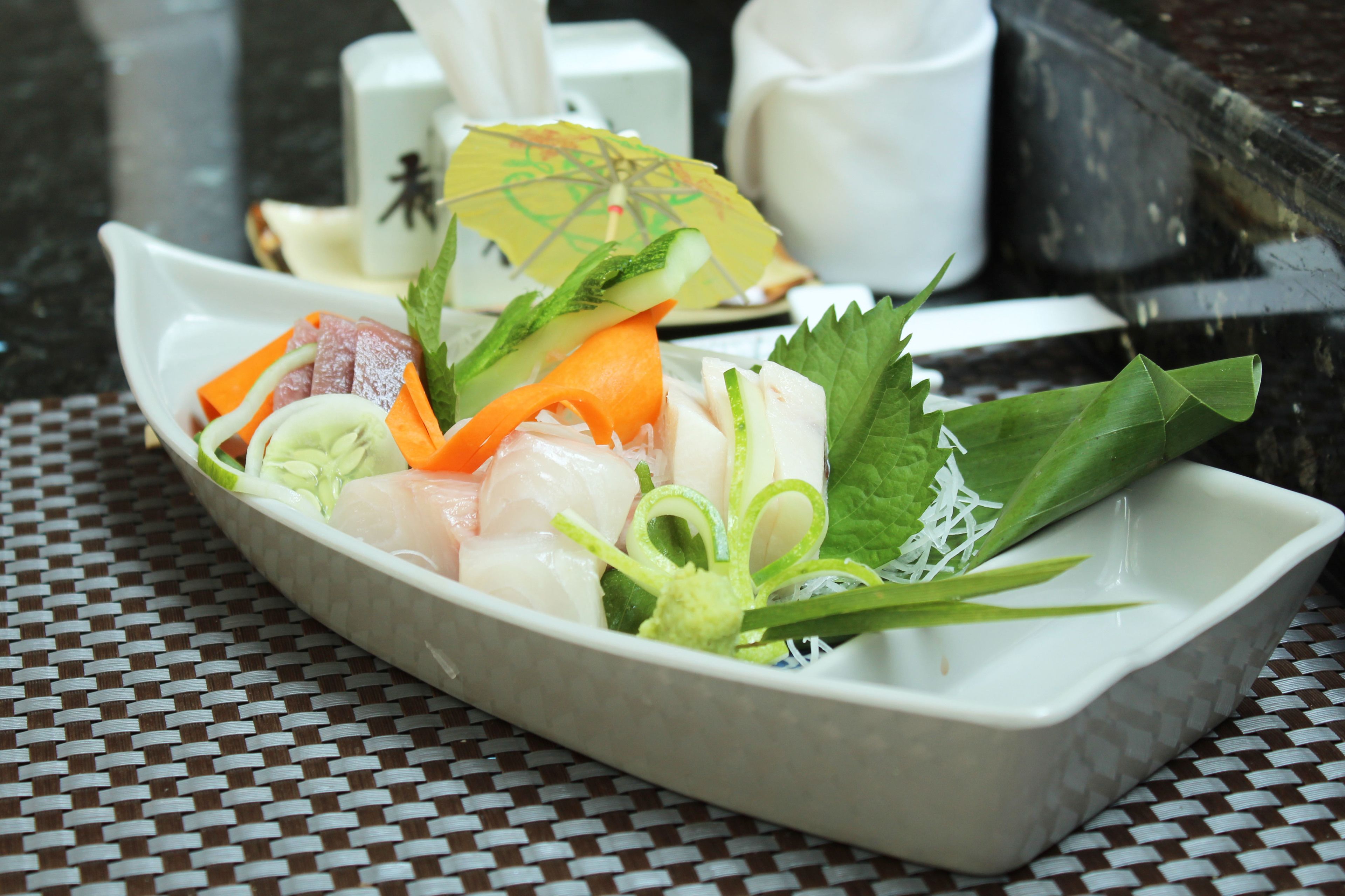 A white rectangular dish with various fresh ingredients, including fish, vegetables, and herbs, on a woven table mat.