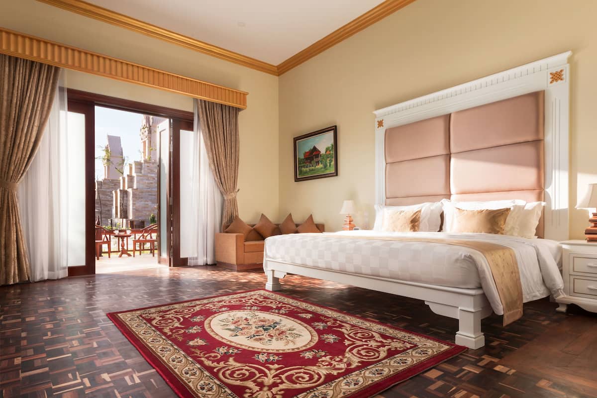 Ein geräumiges Schlafzimmer mit einem großen Bett, einem Teppich mit rotem und goldenem Muster sowie Blick auf ein Gebäude durch eine Glasdoor.