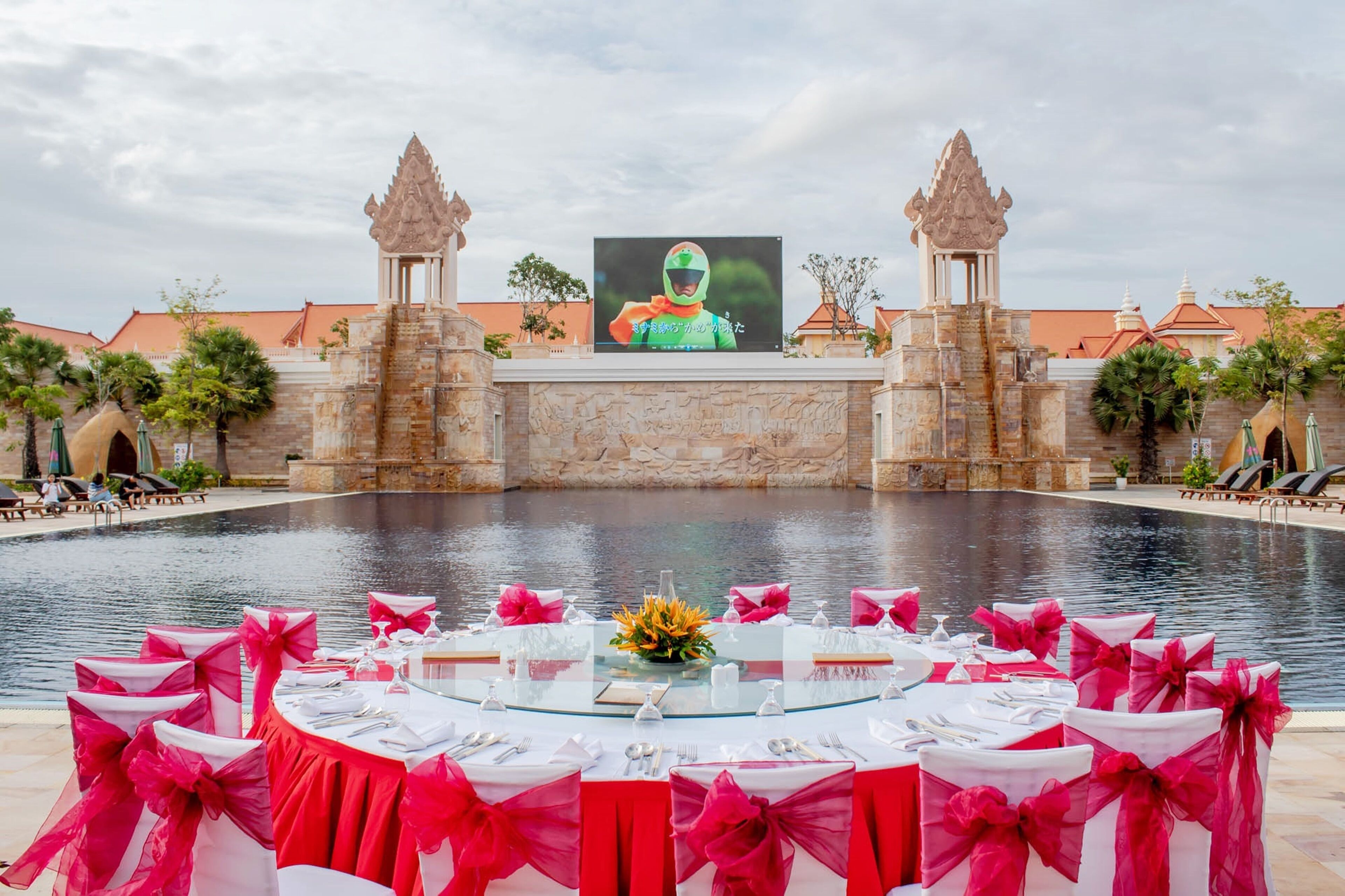 Un área de comedor junto a la piscina con mesas dispuestas para una comida, una pantalla grande mostrando a una persona con casco verde, y arquitectura tradicional al fondo.