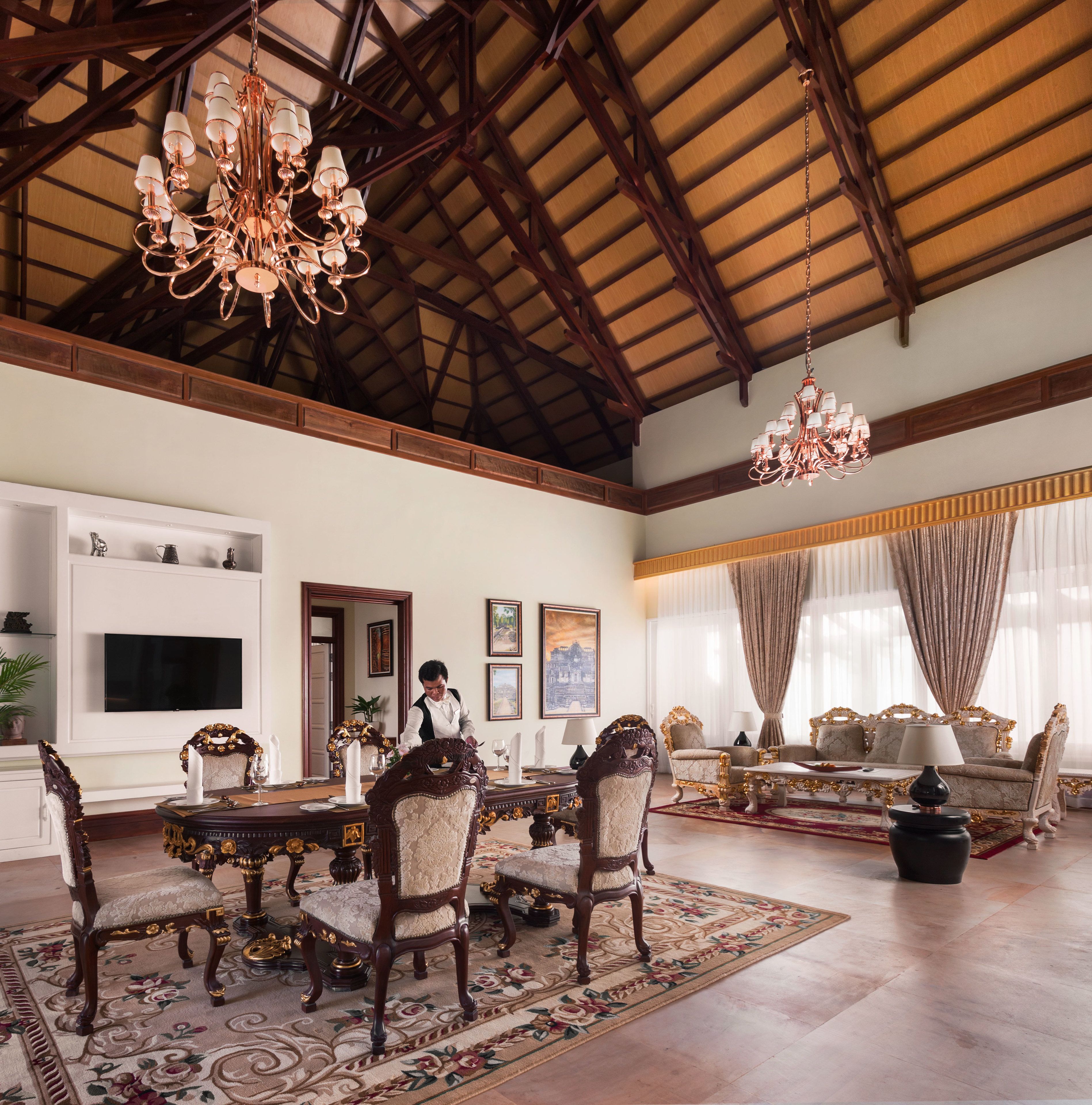 A spacious room with a high ceiling, ornate chandeliers, a dining area with a table set for four, and a seating area with sofas and chairs.