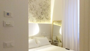 Een hotelkamer met een net opgemaakt bed, een nachtkastje, een spiegel en een aan de muur bevestigde thermostaat.