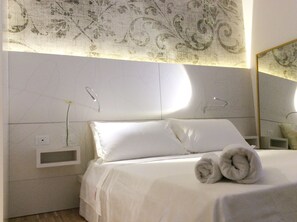 A hotel room with a neatly made bed, a small bedside table with a vase of flowers, and a large mirror on the wall.