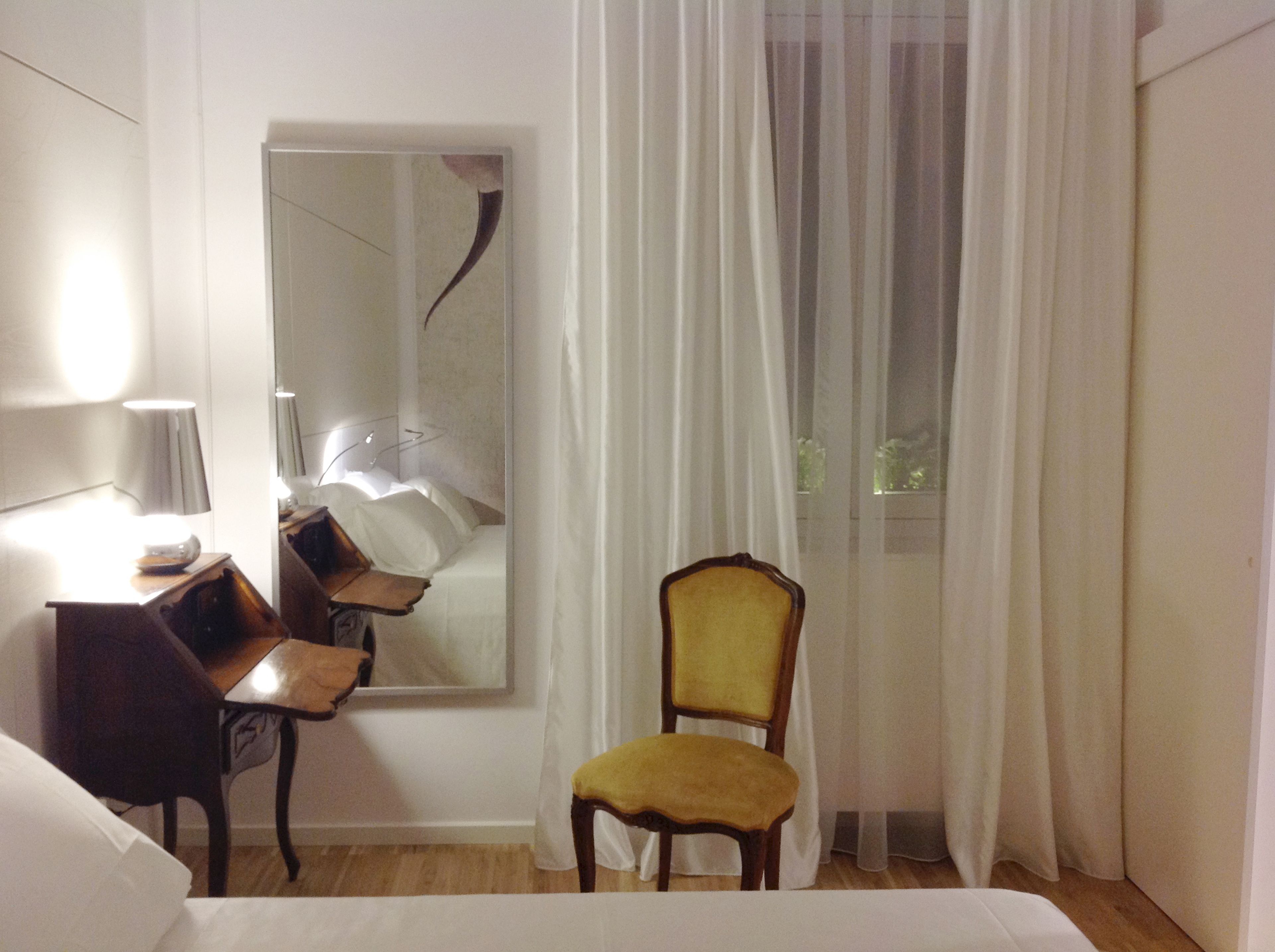 Una habitación de hotel con cama, escritorio con lámpara, silla, espejo y cortinas translúcidas.