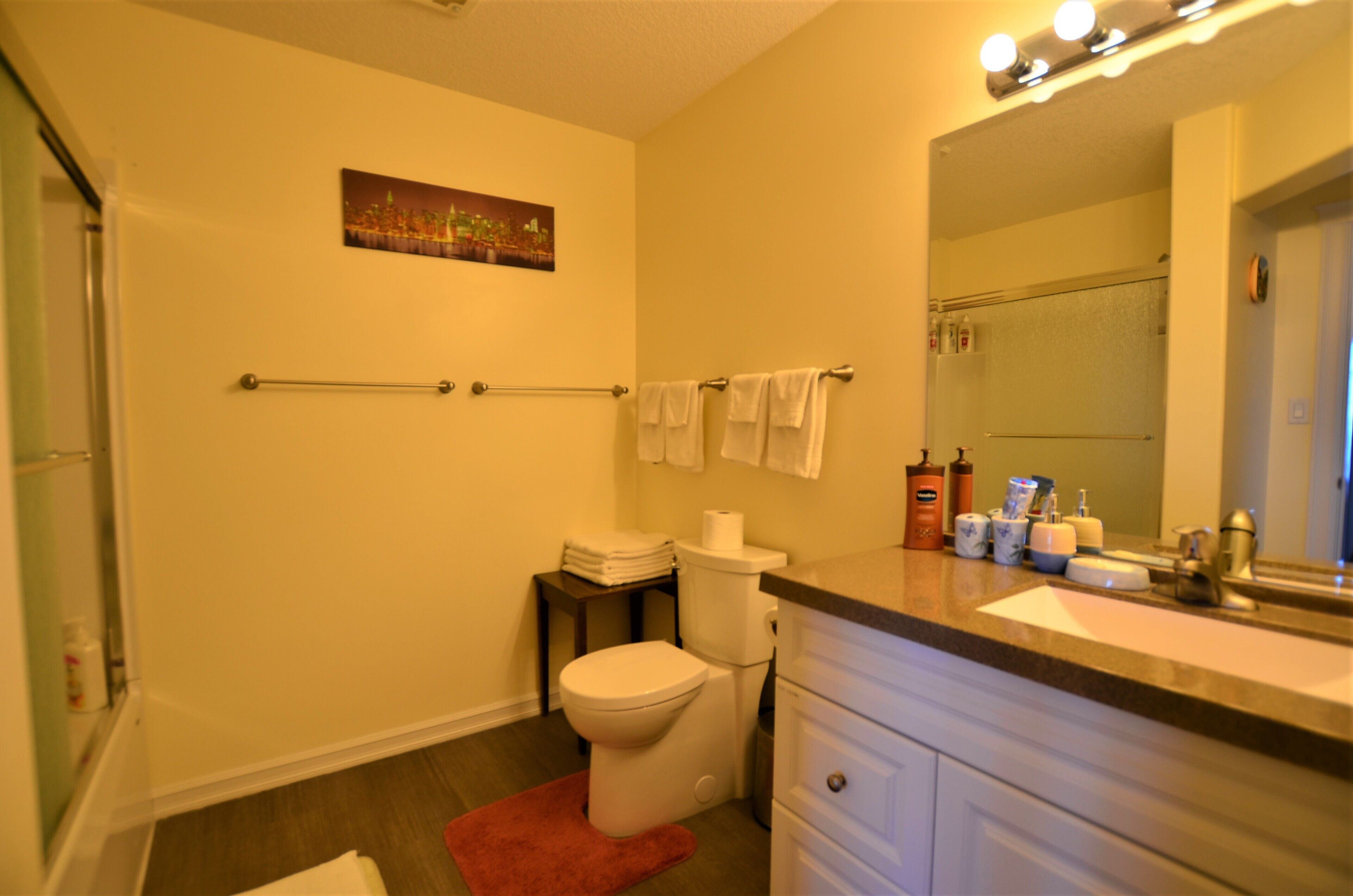 A bathroom with a toilet, sink, and mirror. There is a towel rack with towels, a small table with toiletries, and a painting on the wall.