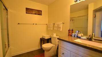 A bathroom with a toilet, sink, and mirror. There is a towel rack with towels, a small table with toiletries, and a painting on the wall.