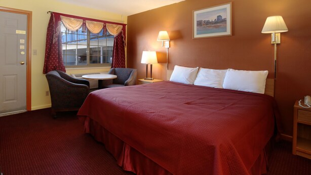 A hotel room with a large bed, a desk with a chair, two bedside lamps, a window with curtains, and a painting on the wall. - Regency Inn (Winnemucca)