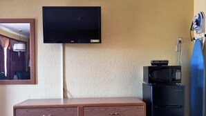 A hotel room with a flat-screen TV mounted on the wall, a wooden dresser with two drawers, a black refrigerator, and a microwave on top of the refrigerator. - Regency Inn (Winnemucca)