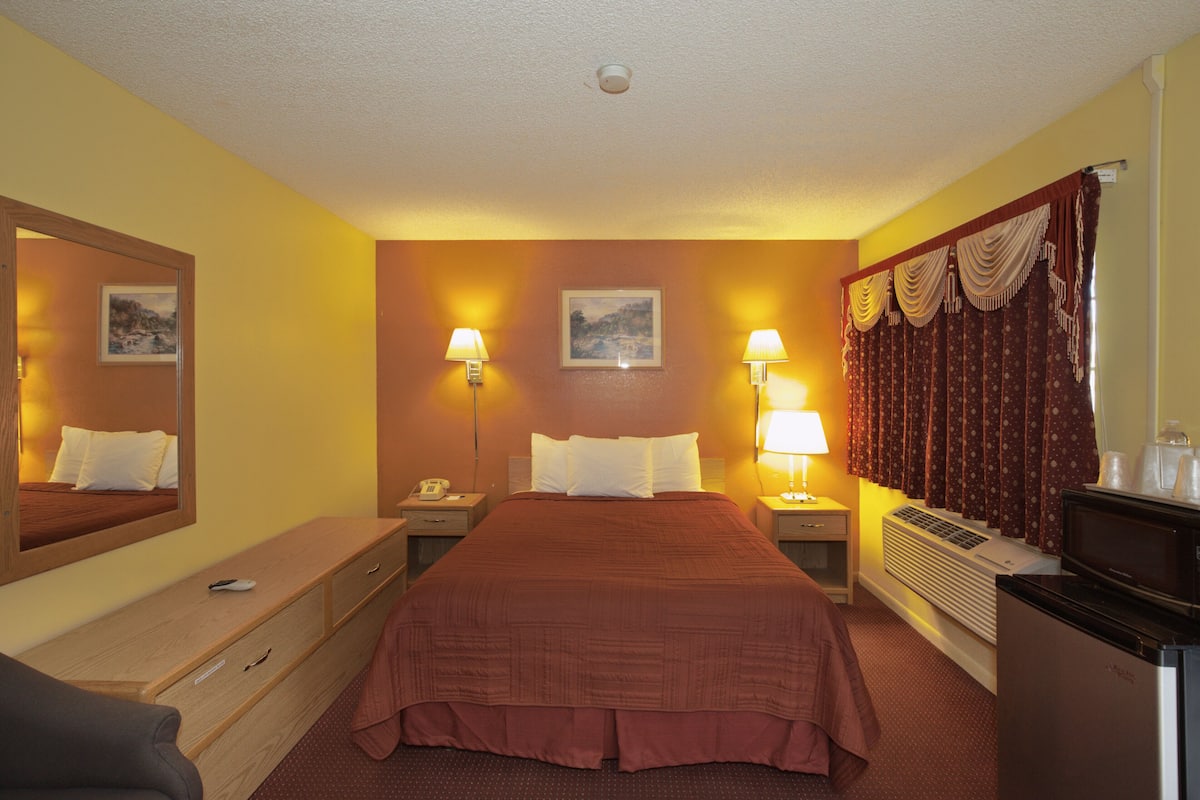 A hotel room with a large bed, two bedside tables with lamps, a dresser, a desk, a mirror, and a refrigerator.