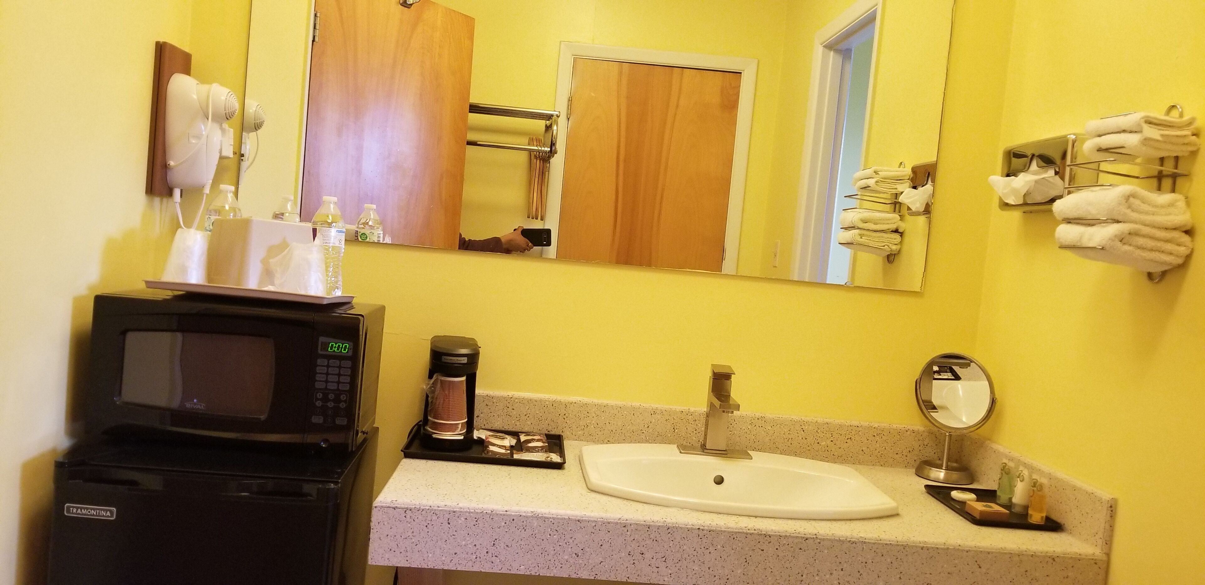 A bathroom with a sink, mirror, towel rack, hairdryer, and coffee maker.