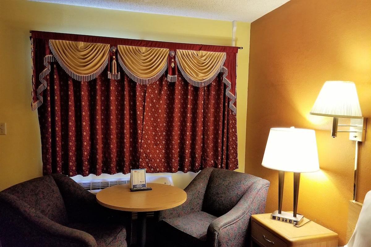 A hotel room with a bed, two armchairs, a small table, a lamp, and a window with curtains.