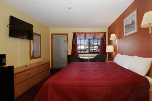 A hotel room with a large bed, a TV mounted on the wall, a desk with a chair, a window with curtains, and a lamp on the nightstand.