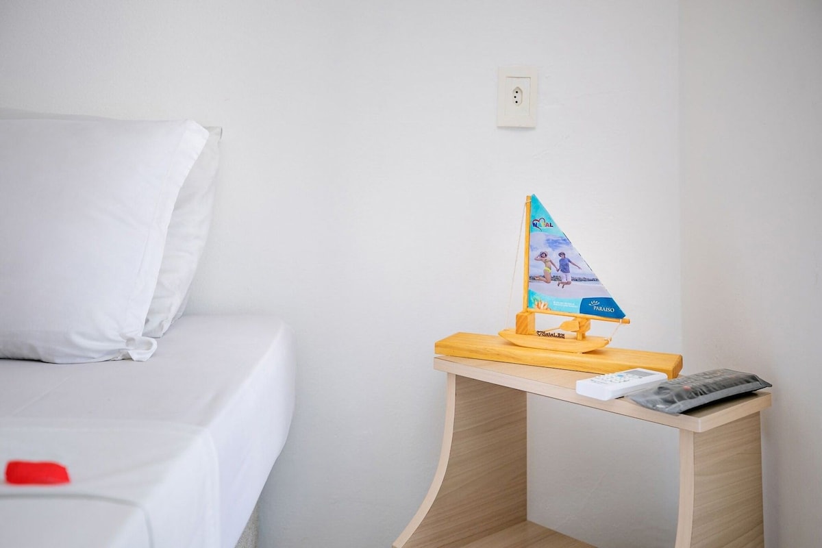 A neatly made bed with white bedding, a small wooden nightstand with a model sailboat, and a remote control on it.
