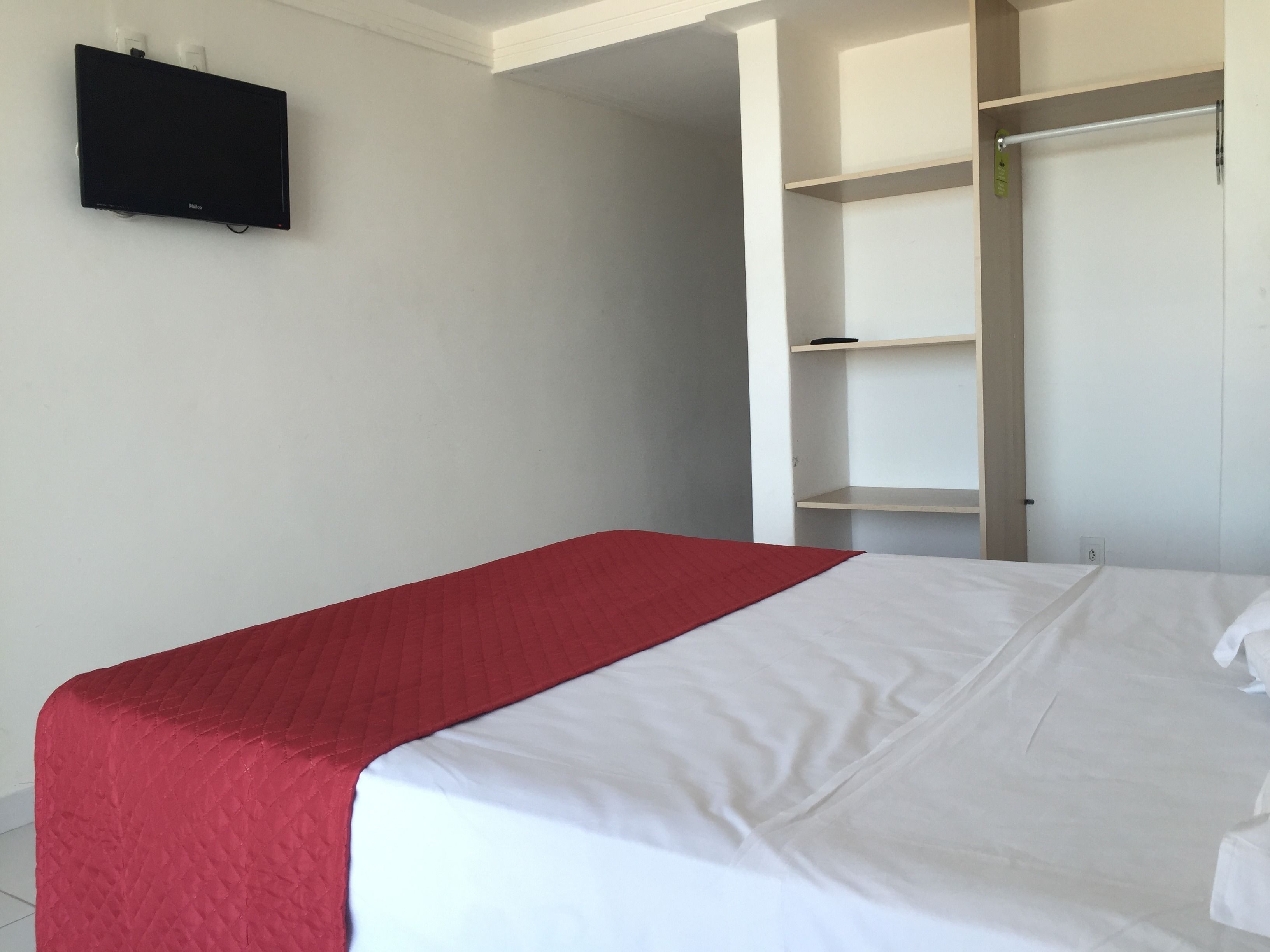 Une chambre à coucher avec un lit, une couverture rouge, une couette blanche, un téléviseur fixé au mur et une armoire avec un miroir.