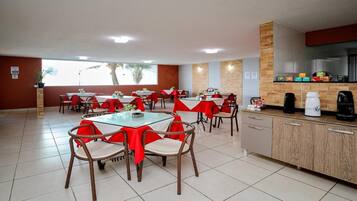 Een restaurant met ronde tafels en rieten stoelen, rode tafelkleden en een keuken met koffieautomaten.