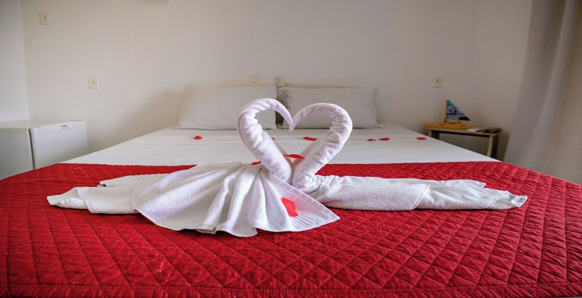 Habitación de hotel con una cama cubierta por una colcha acolchada roja y una toalla blanca doblada en forma de dos cisnes.